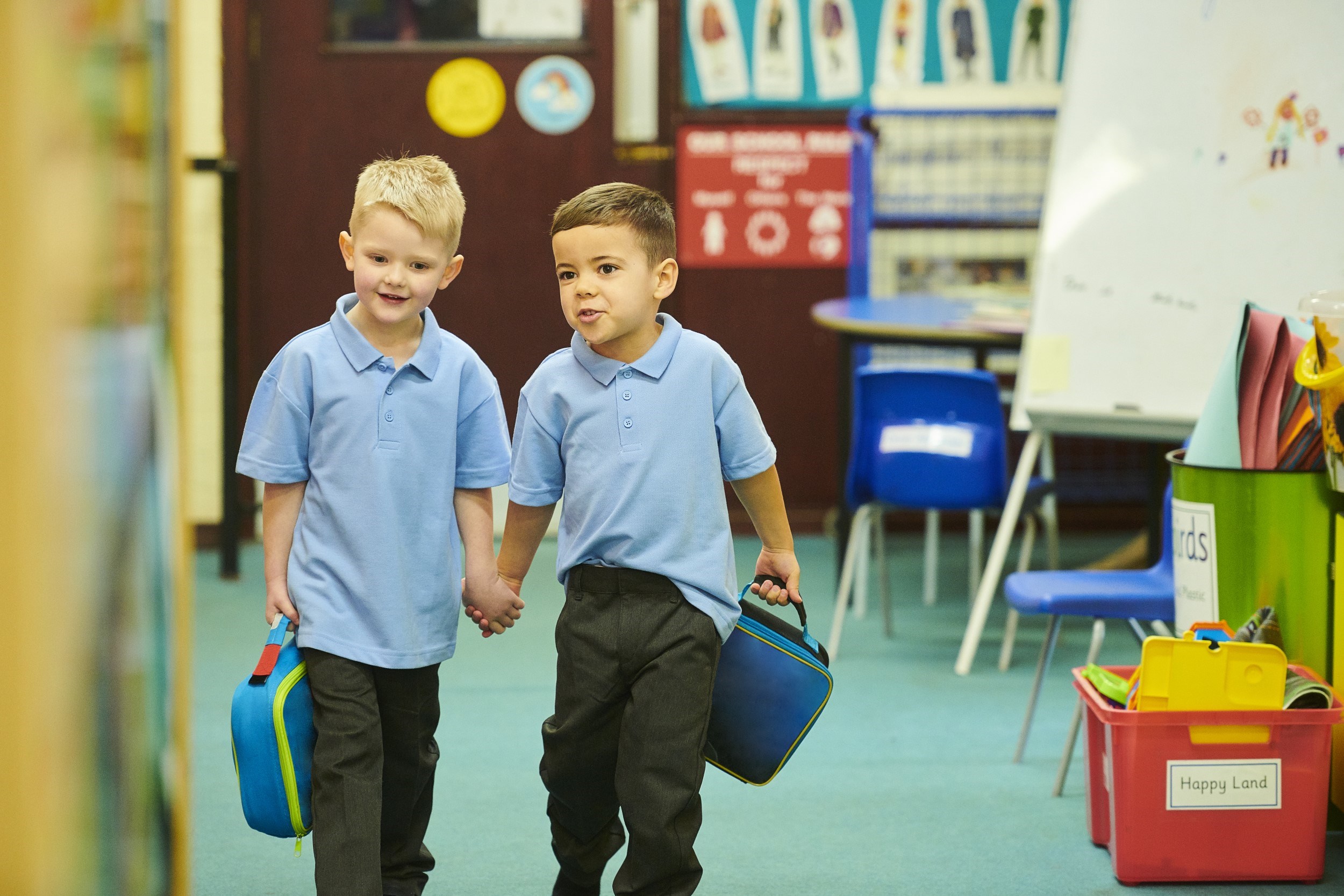 Two children holding hands and their bags walking through a classroom
