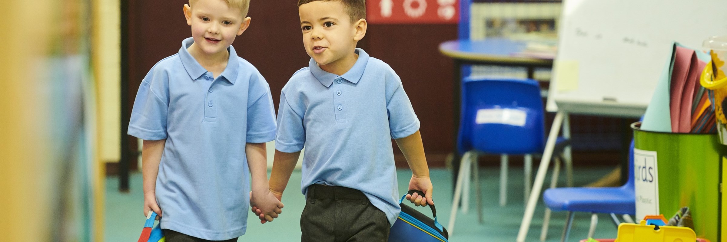 Two children holding hands and their bags walking through a classroom