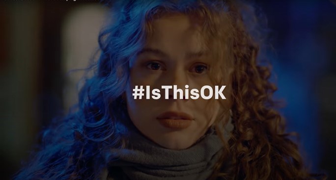 A girl's face looking straight ahead whilst on a night out, with glowing lights in the background and with the hashtag #IsThisOK written in white letters over the top of the image.