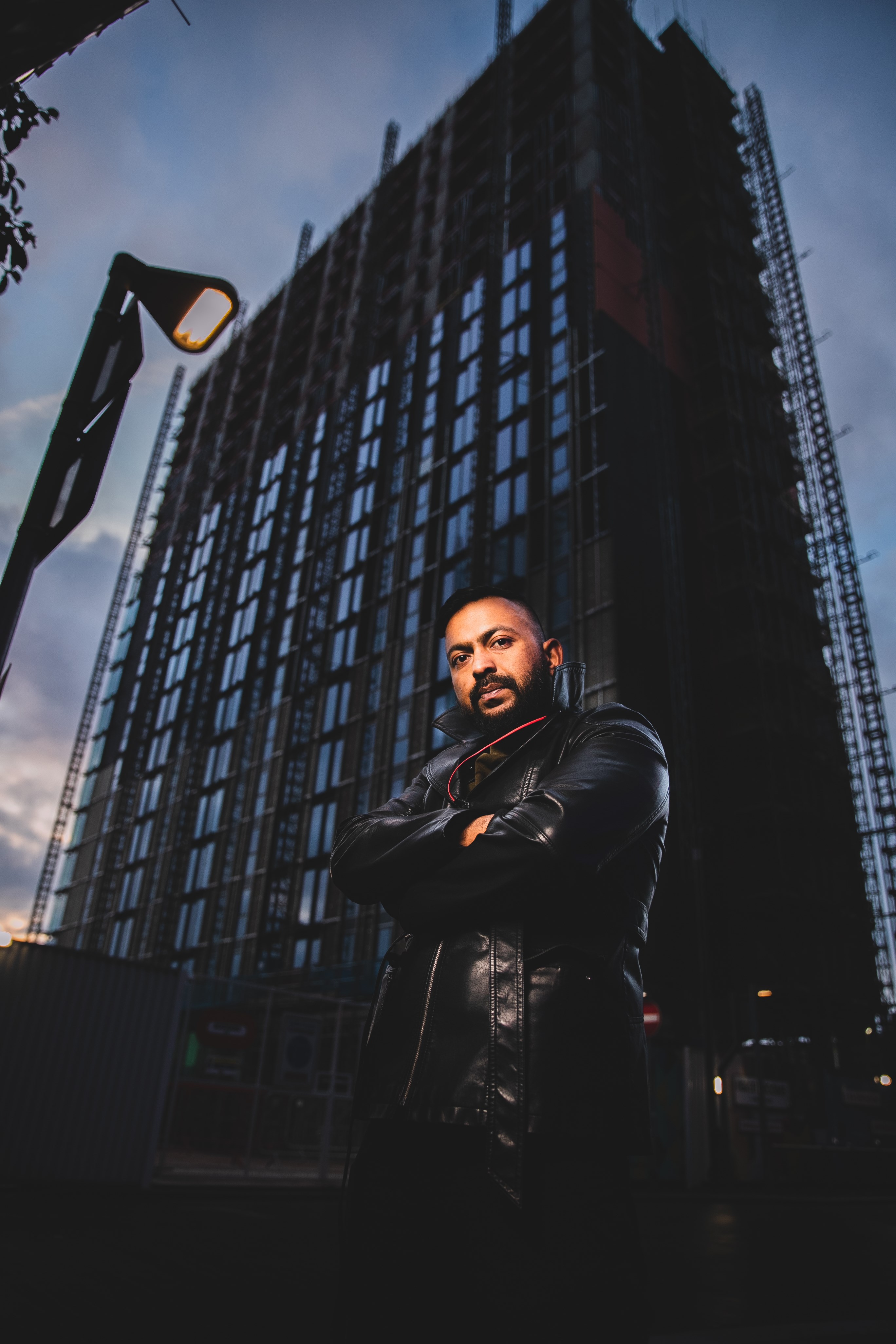 Photo of Sam Malik with the camera low down looking up at him with a high rise office block behind him, lit in a dramatic dark way. 
