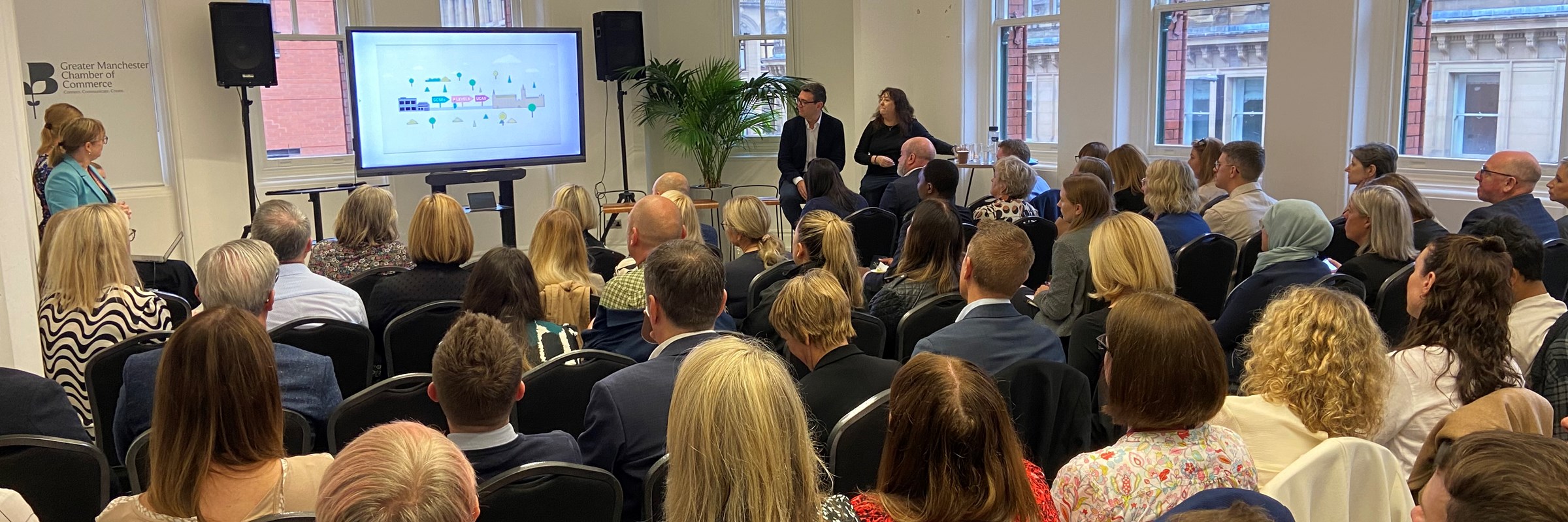 Andy Burnham stands by a presentation screen in front of a full audience at a business breakfast held at GM Chamber of Commerce
