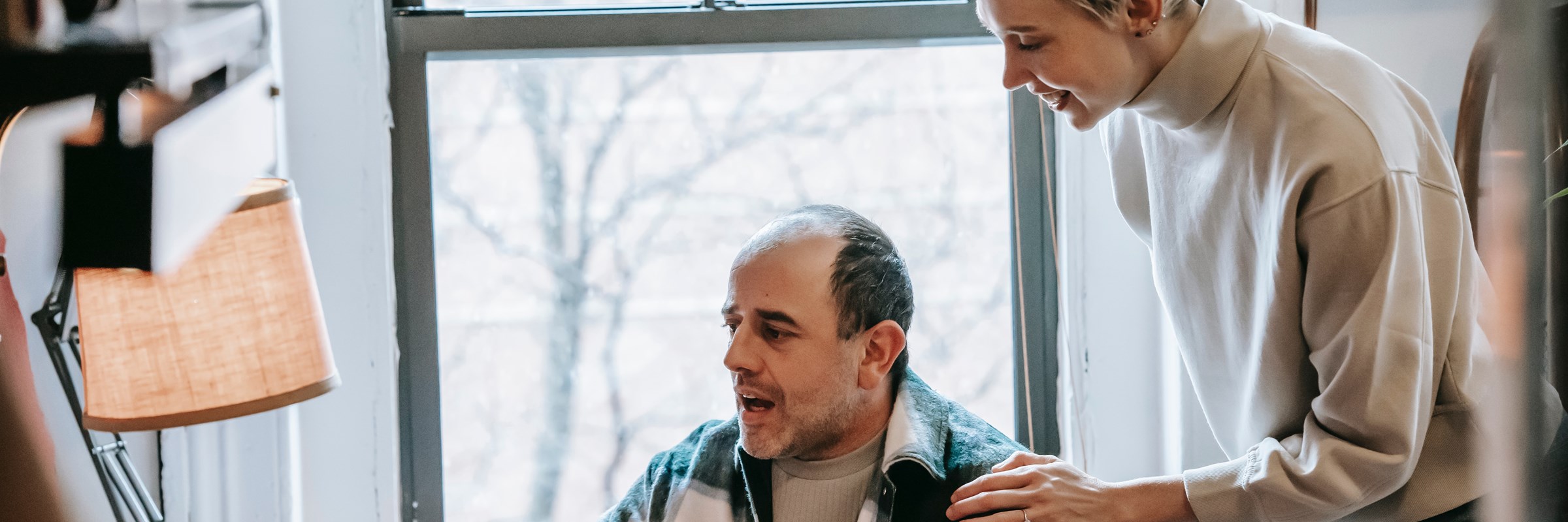 A middle aged man is using a laptop. A younger woman stands over him with her hand on his shoulder as though she is supporting him with how to use it