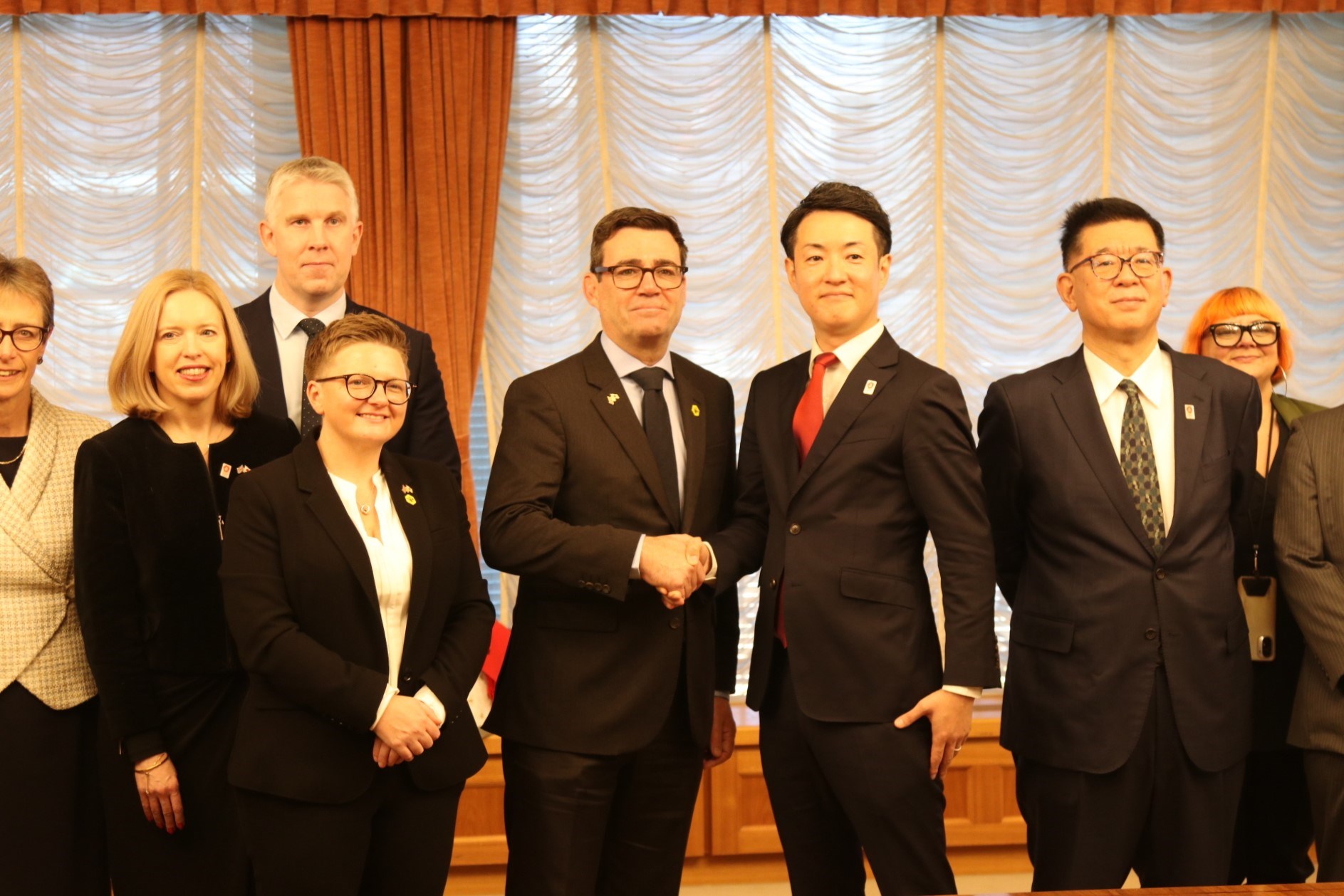 Andy Burnham and Mayor Hideyuki Yokoyama stand side-by-side, shaking hands, flanked by other members of the Greater Manchester delegation