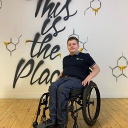 An image of a young man in a wheelchair in the Federation House building next to a wall saying This is the Place