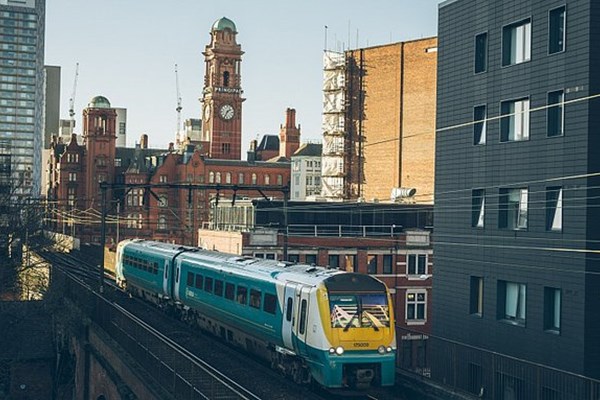 Teal and yellow train on railway bridge. In the background you can see a tall tower of an old building and other more modern buildings surround the train.
