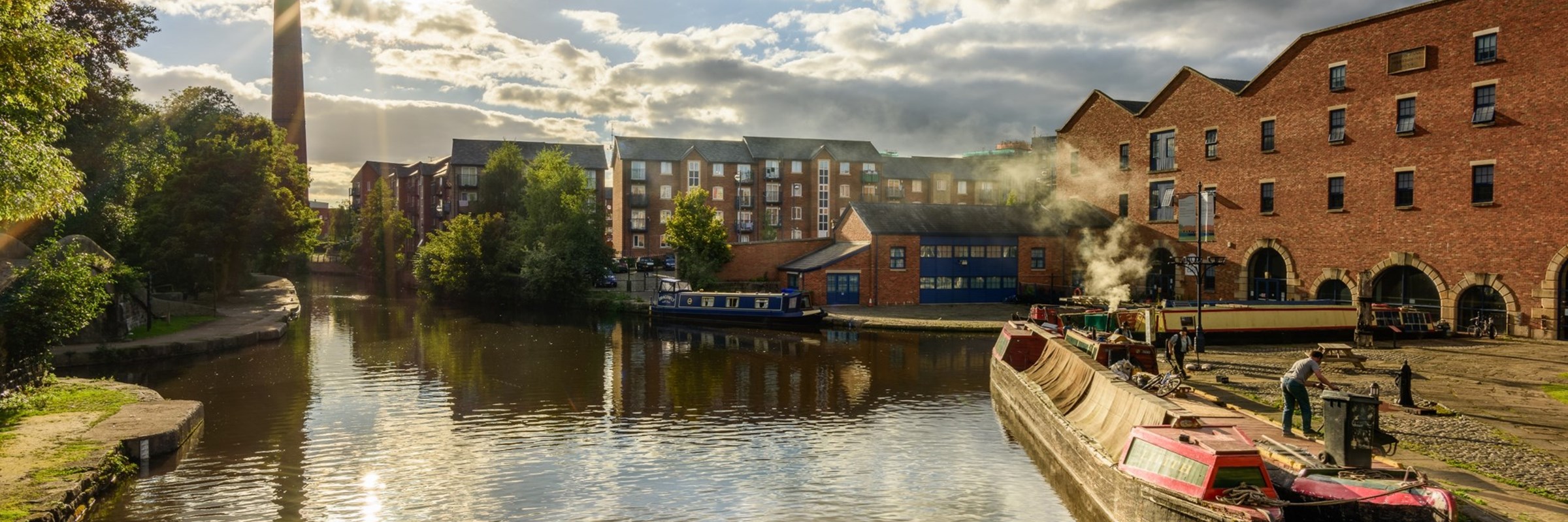 A canal barge parked at a dock, with buildings and a chimney in the distance.