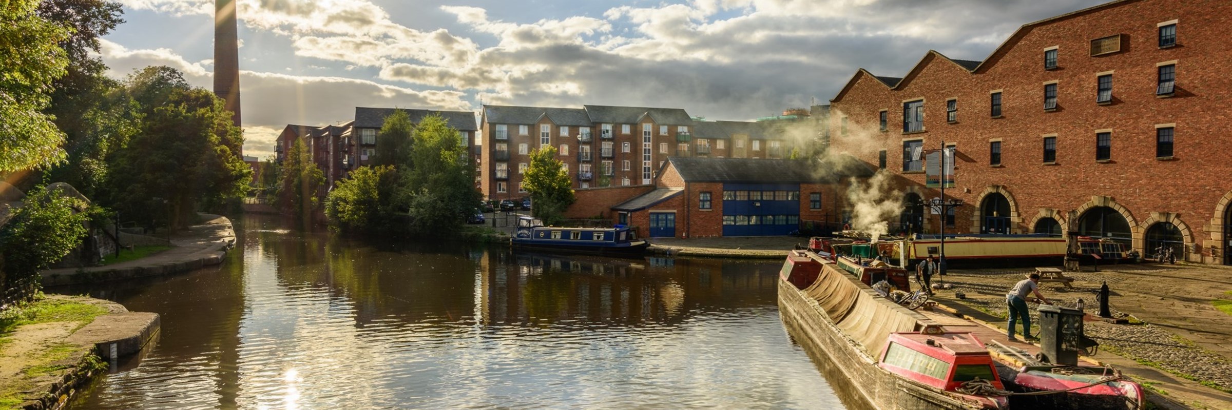 A canal barge parked at a dock, with buildings and a chimney in the distance