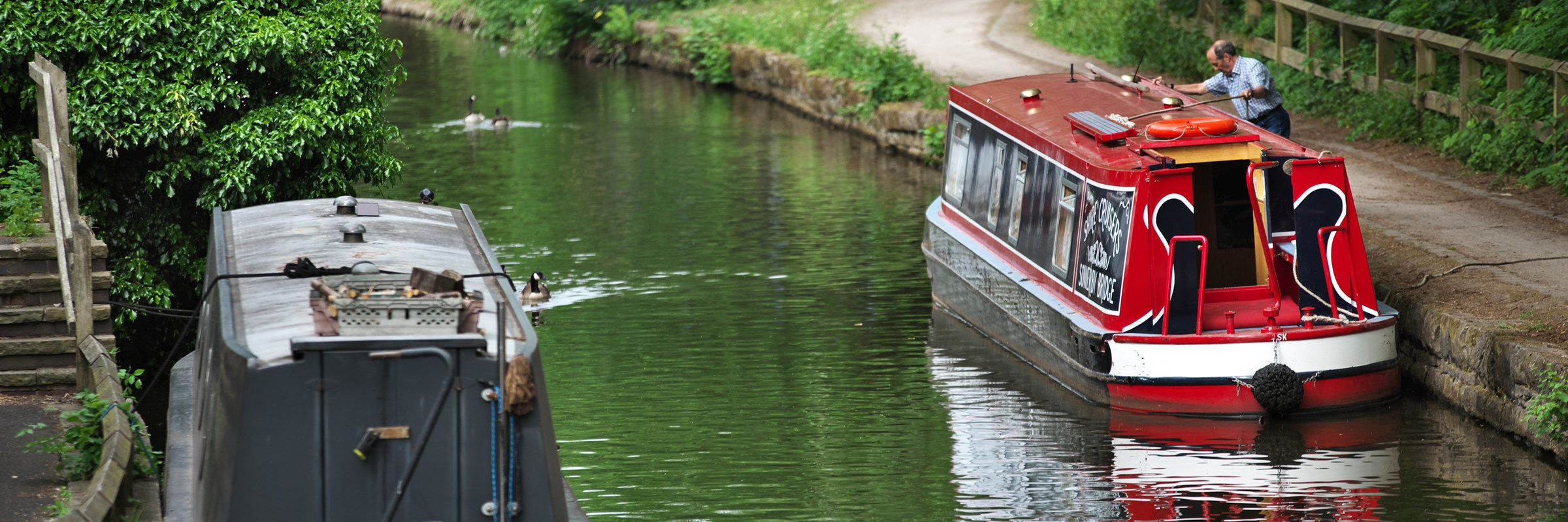 Two docked canal boats situated in the canal water beside a pathway, and surrounded by tree area.