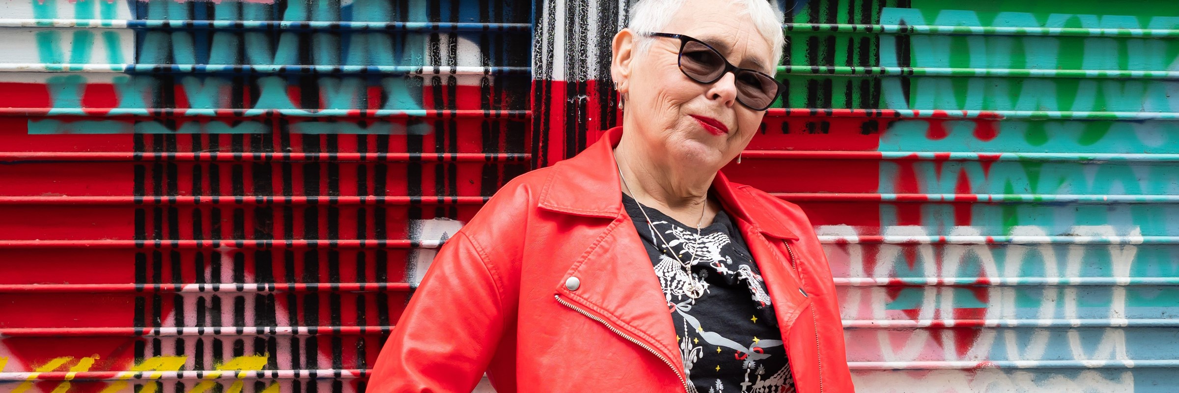 An older person wearing sunglasses standing in front of a multicoloured background.