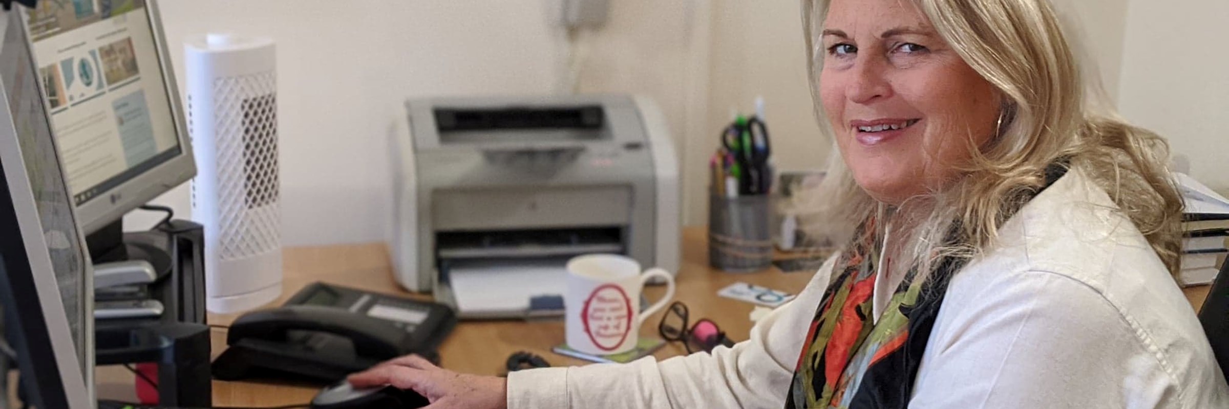 Ladderstore MD Gail Hounslea sits at a desk in front of a keyboard and monitor - she has a light jacket, coloured scarf and blonde hair