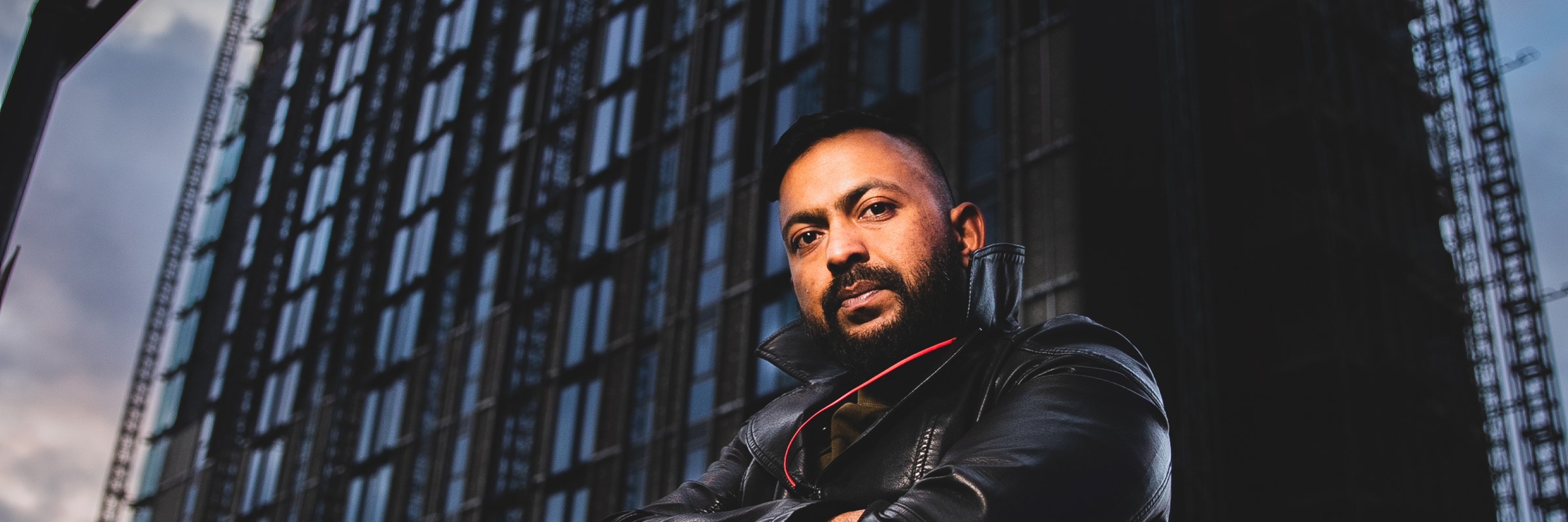 Photo of Sam Malik with the camera low down looking up at him with a high rise office block behind him, lit in a dramatic dark way.