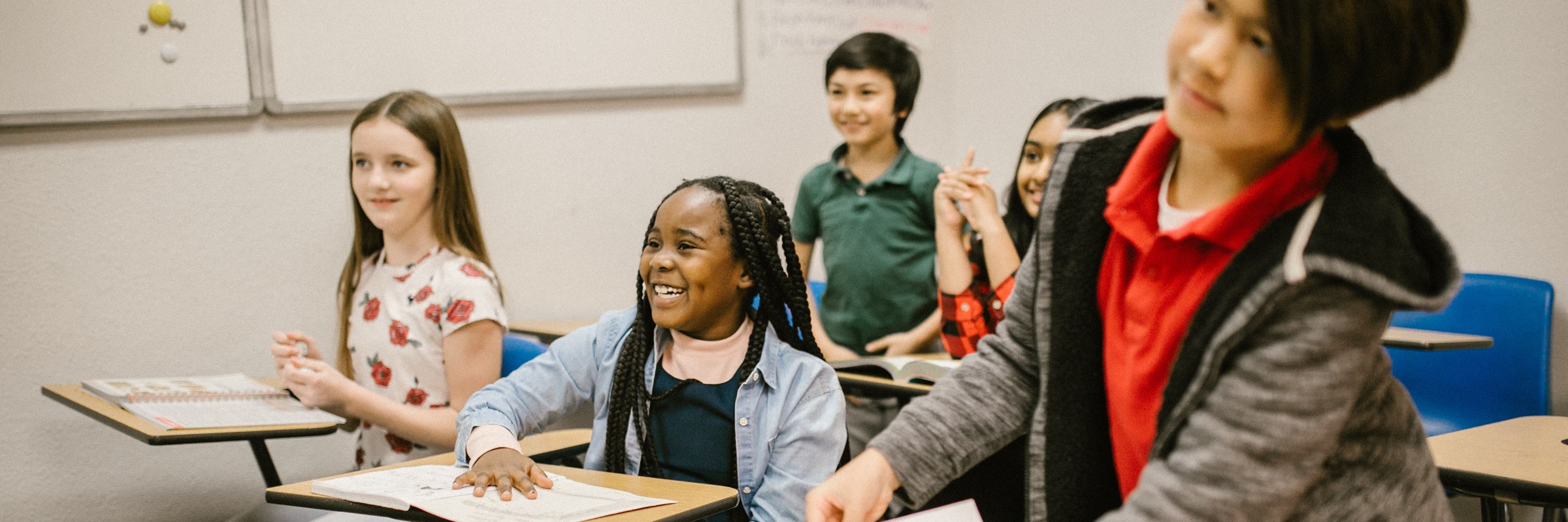 Young children in a classroom, they are smiling and sitting on individual desks with papers in front of them