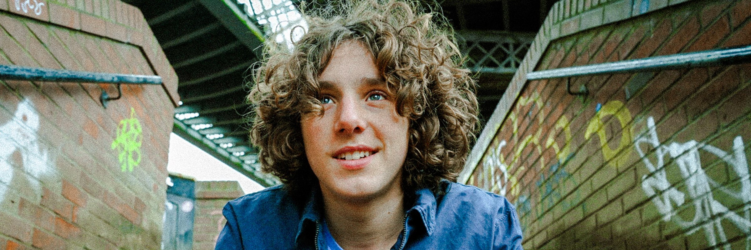 Image of Alex Spencer in a blue top and Jacket sat on some steps with his hands clasped together. There is some graffiti on the walls of the steps to the side of him.