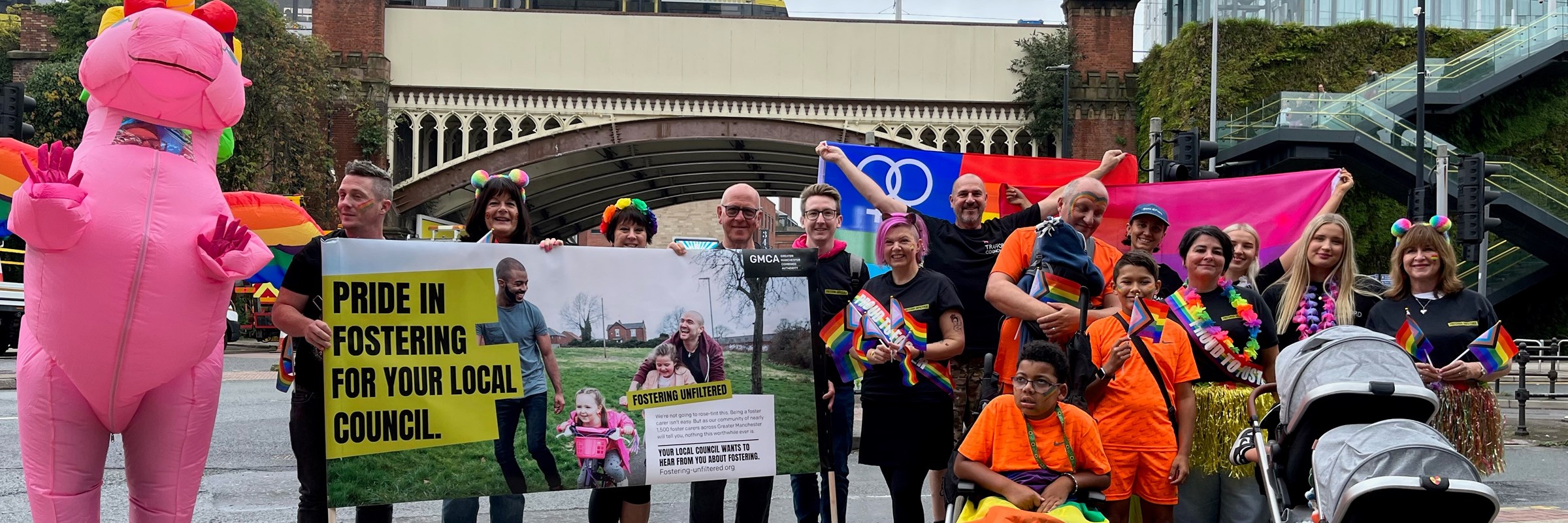 Group shot of adults and children with a banner for the campaign that says 'Pride in fostering for your local council'. Everyone is dressed in pride themed items and one is dressed in a blow up pink unicorn outfit.