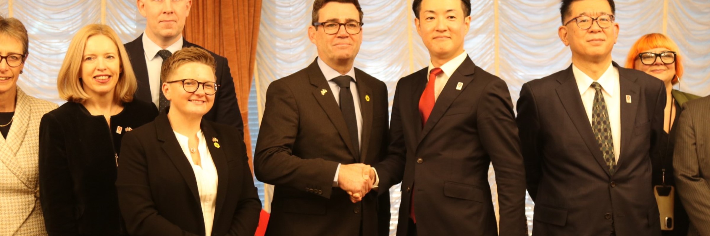 Andy Burnham and Mayor Hideyuki Yokoyama stand side-by-side, shaking hands, flanked by other members of the Greater Manchester delegation