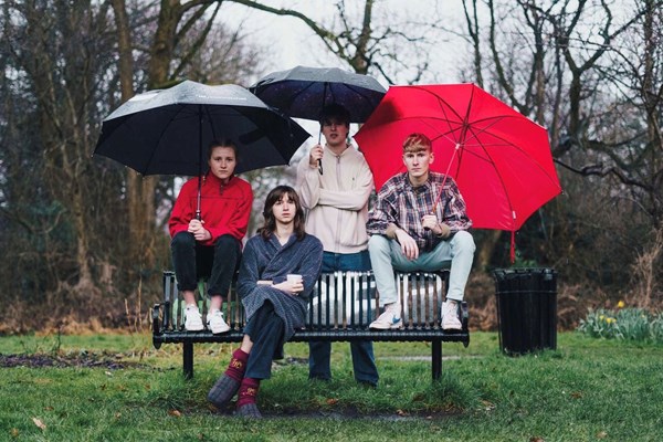 Image of the 4 band members in a park sat on a bench with three umbrellas over them