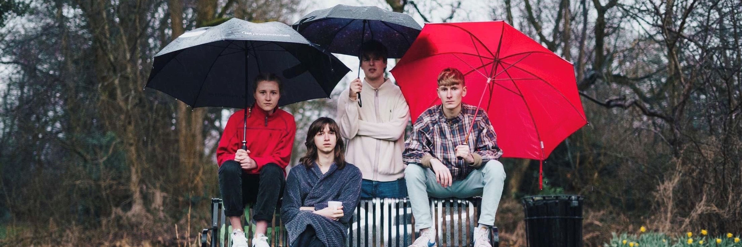 Image of the 4 band members in a park sat on a bench with three umbrellas over them