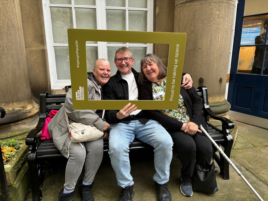 Three people huddles together, holding a cardboard picture frame and smiling
