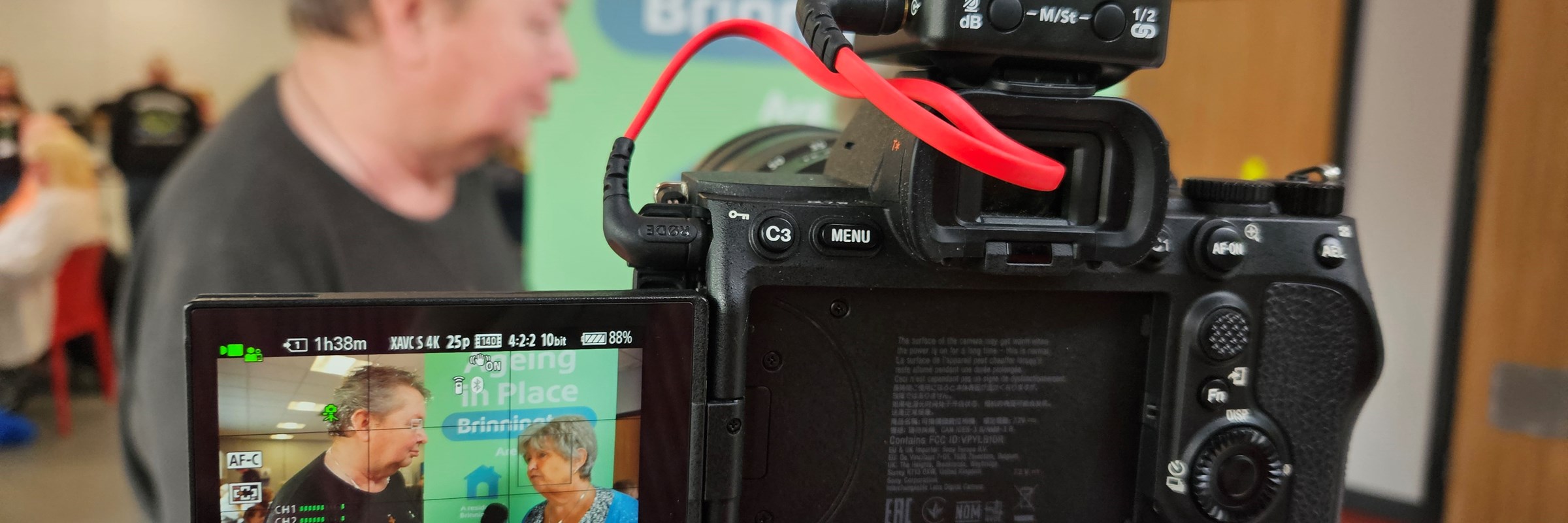 A camera screen displays an interview in progress with two people, while one of them stands blurred in the background.